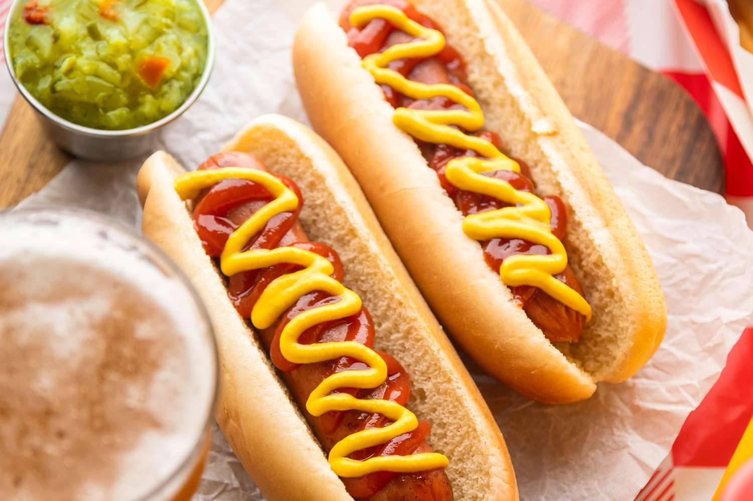 Two hot dogs with mustard and ketchup, served on a wooden board with a side of relish and a drink visible