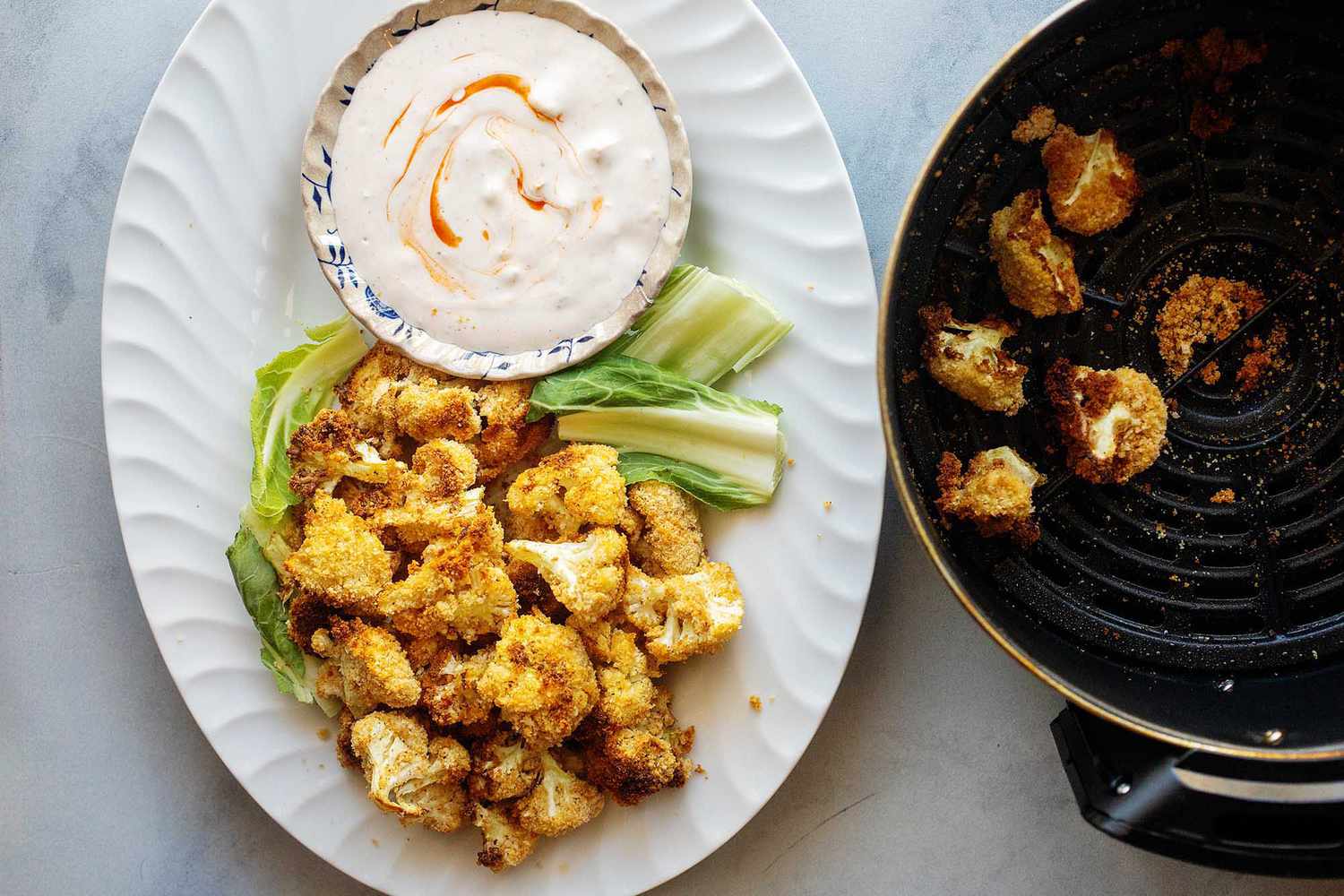 Hortizontal photo with a white fluted platter sitting vertically on a marble background. The platter has golden air fried cauliflower heaped on the bottom with a few leaves of lettuce and above is a small bowl of creamy dip swirled with hot sauce. To the right of the platter is the air fryer with a few fried cauliflower inside.