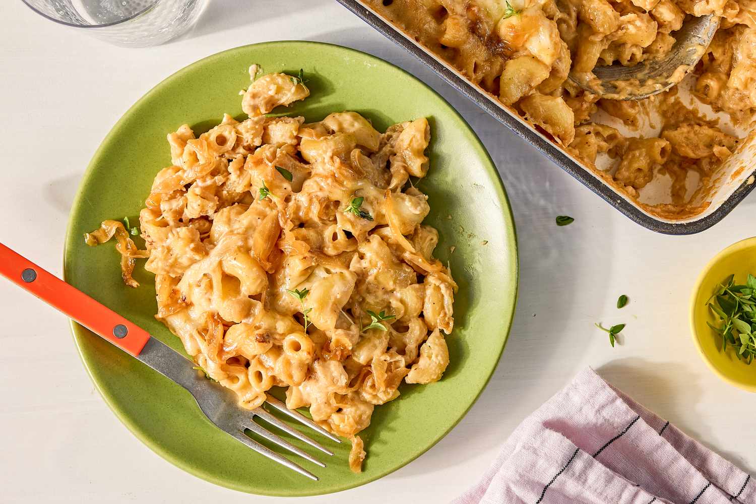 A plate of French onion mac and cheese served with a fork nearby is a baking dish containing more of the dish