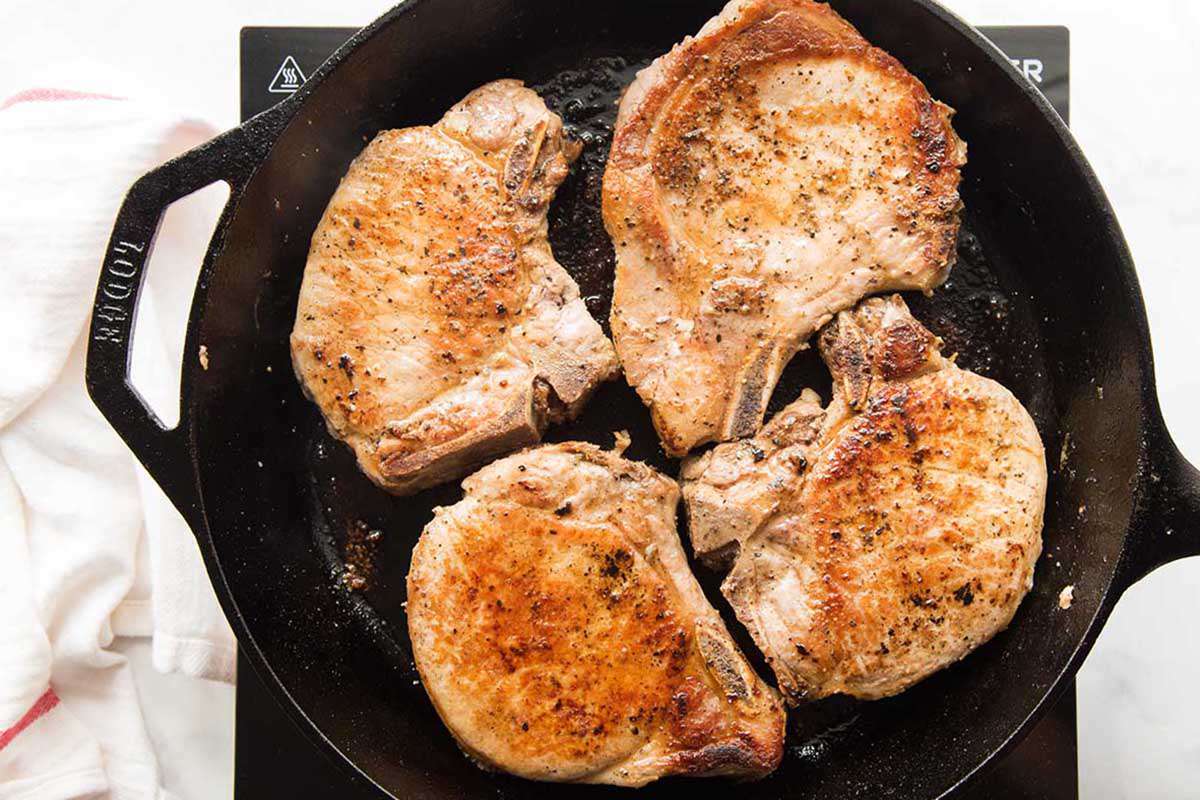 Pork chops browned in a cast iron skillet with a hand sprinkling salt and pepper over the chops.
