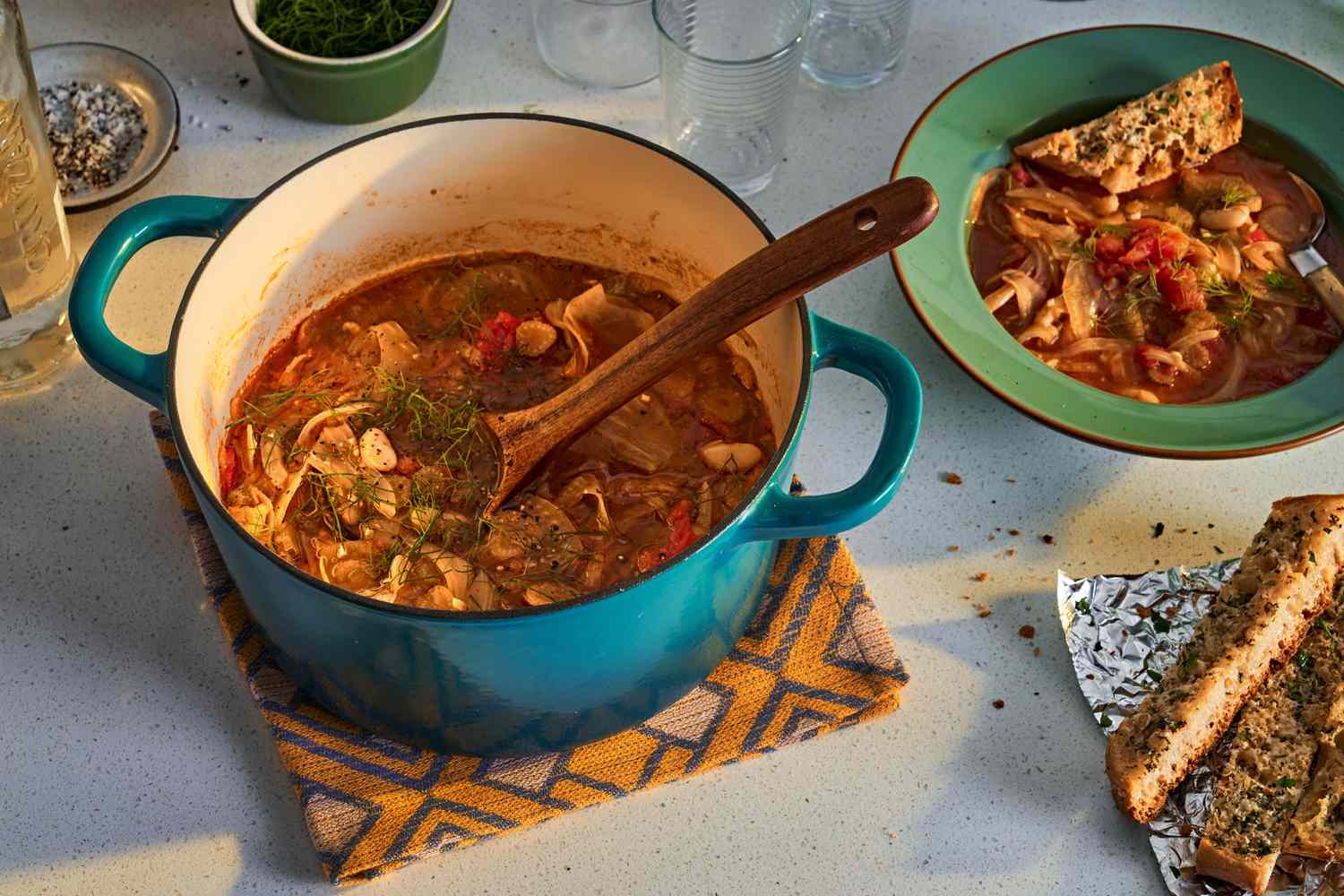 Dutch oven filled with tomato, white bean, and fennel stew on a kitchen counter, a bowl of the soup and garlic bread alongside