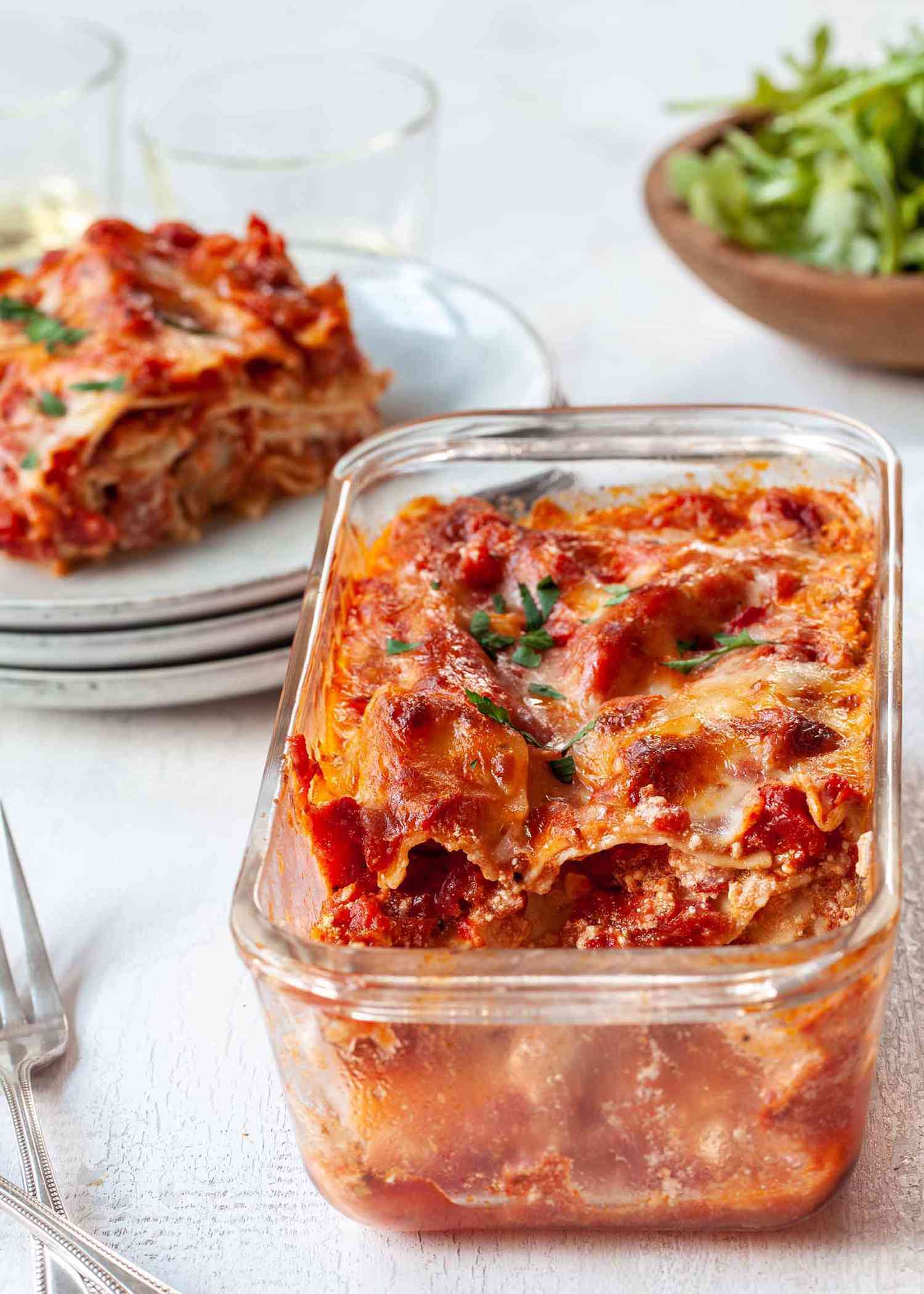 Vertical view of small batch lasagna with a piece missing inside the glass loaf pan. Behind is a stack of white plates with the slice of lasagna on top. Two tumblers and a small bowl of lettuce are visible at the top of the image.