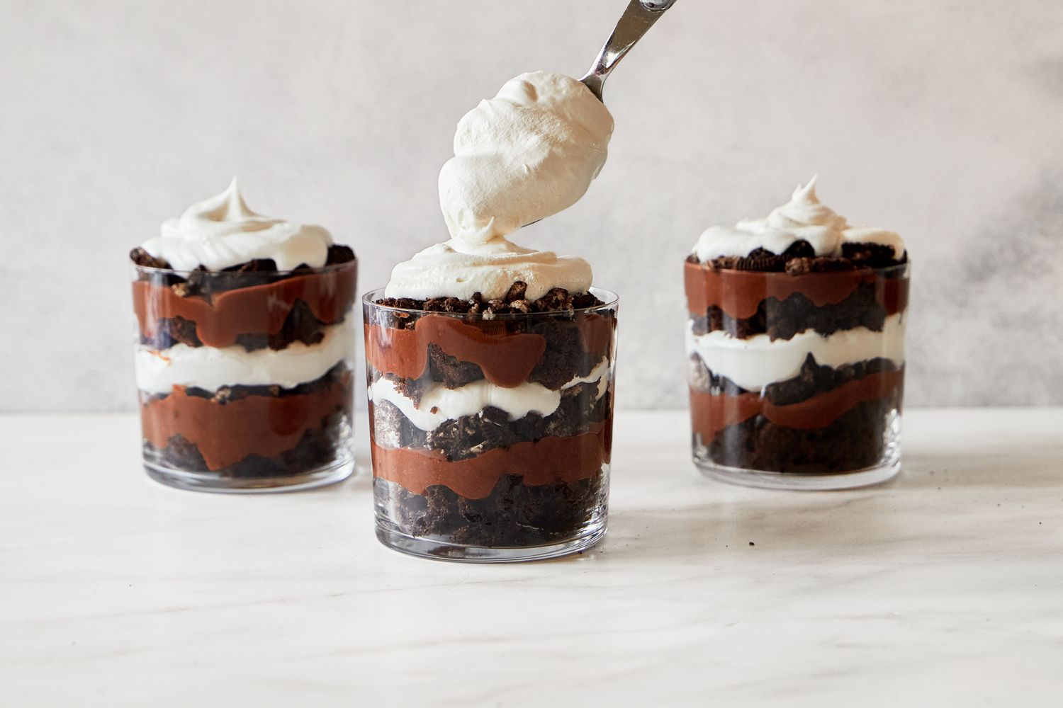 Three servings of layered chocolate and cream dessert in glass cups with whipped cream being added to one