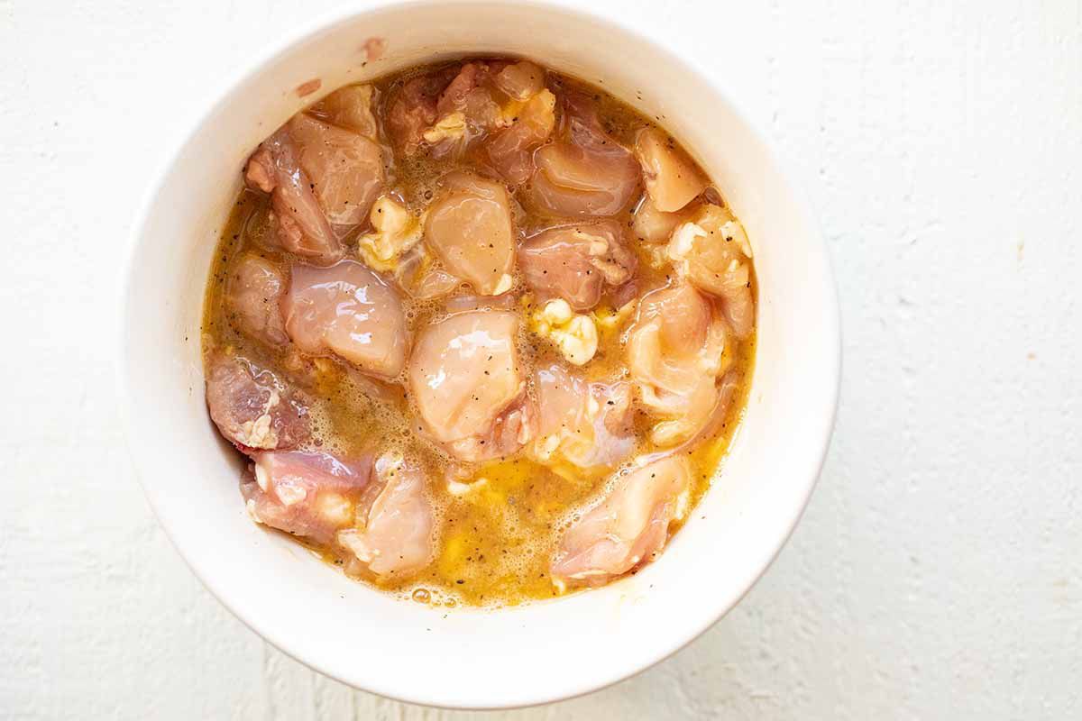 Whisked eggs and raw chopped chicken mixed together in a white bowl to make take out chinese orange chicken.