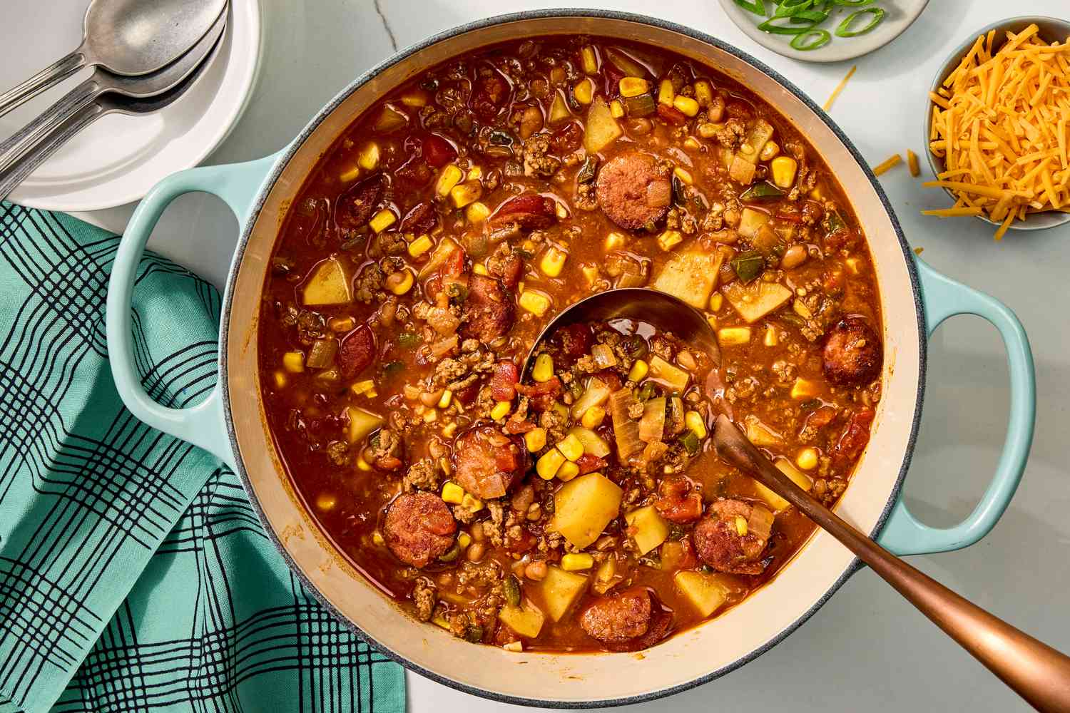 Overhead view of a dutch oven of cowboy stew with a ladle next to a bowl of shredded cheddar, scallions, spoons, and a cloth napkin all on a marble countertop