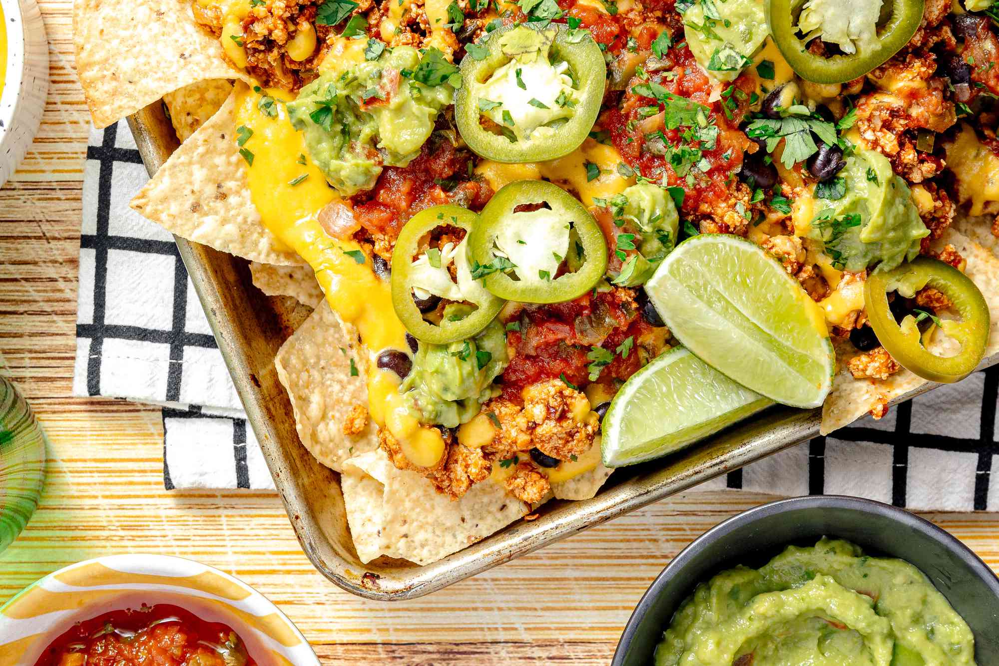 Baking Sheet of Vegan Nachos (Nachos Topped with Salsa, Queso, Pickled Jalpenos, and Two Lime Wedges) Surrounded by Bowls of Guacamole, Salsa, and Queso
