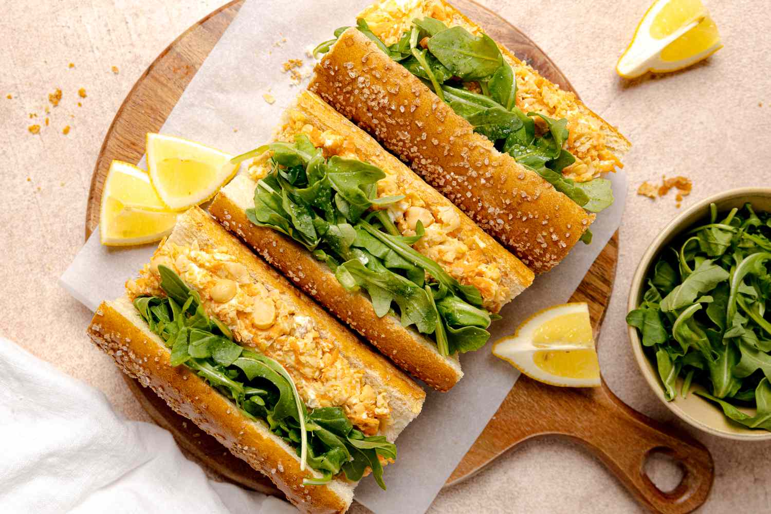 Chickpea Paninis on Parchment Paper over a Circular Wooden Board Surrounded by Lemon Wedges and a Bowl of Arugula