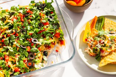 A glass dish containing layered nacho dip topped with ingredients such as olives herbs and vegetables served with tortilla chips on a plate