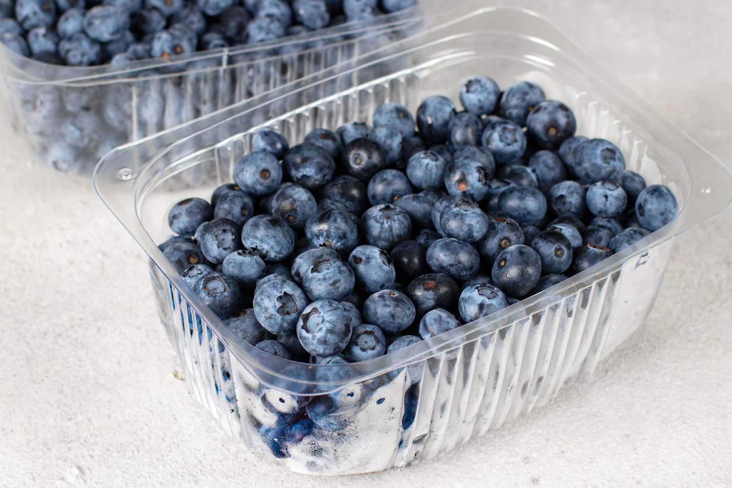 Two clear plastic containers of blueberries with no lids on a white surface