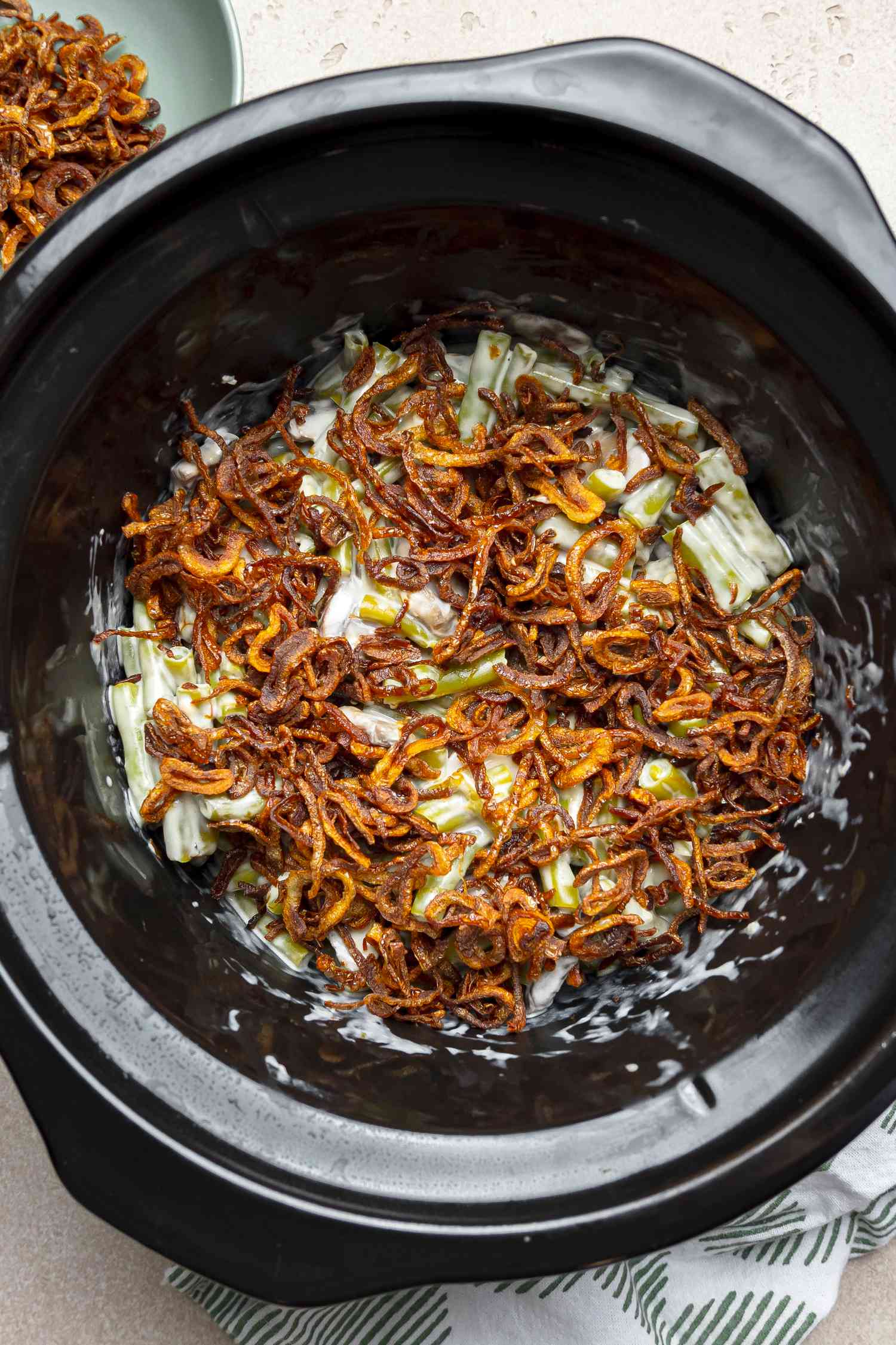 Green Bean Casserole in a Slow Cooker Topped with Crispy Shallots and Next to It, a Small Plate with More Shallots and a Kitchen Towel