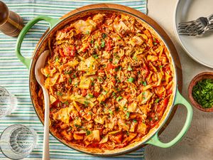 unstuffed cabbage rolls in a large pan at a table setting with a stack of plates, a small bowl of minced herbs, glasses of water, and a pepper cracker