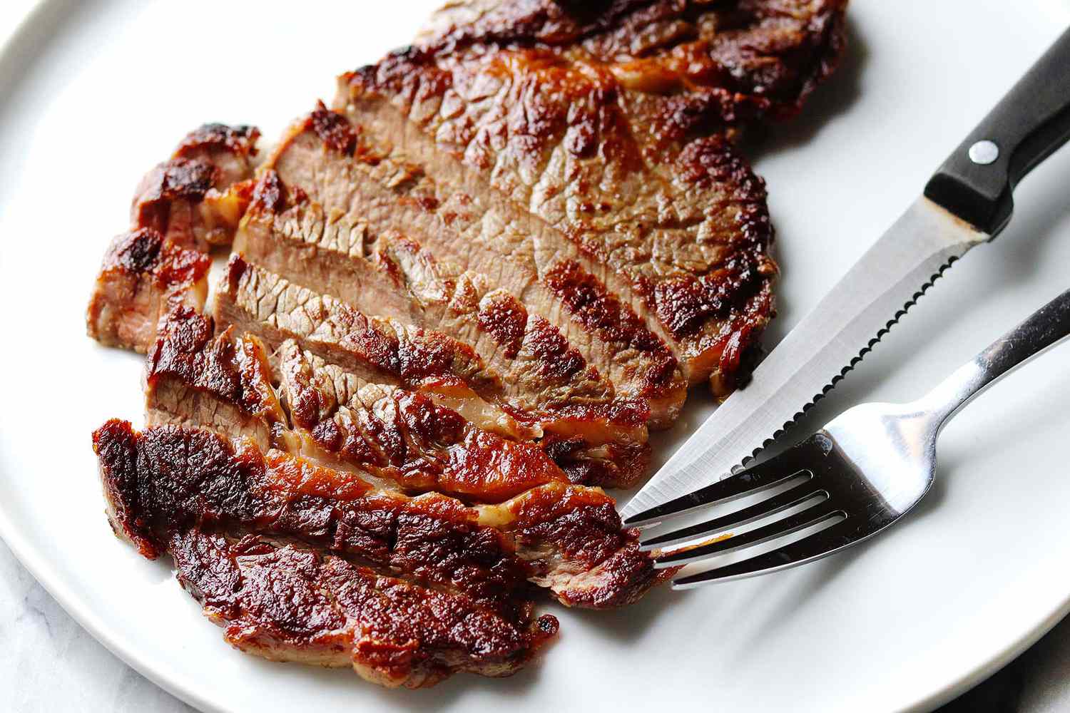 Bone-in steak with a dark crust from being cooked on a stove. It is sliced in half inch pieces horizonally. A steak knife and fork rest to the right of the steak.