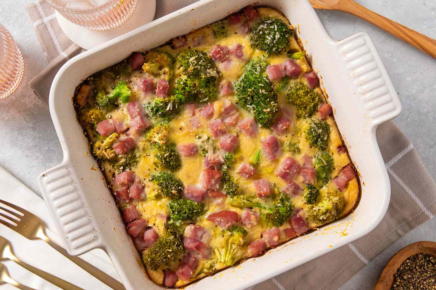 Overhead view of a white casserole dish of broccoli cheese pie in a table setting with forks, glasses and a bowl of pepper to the side