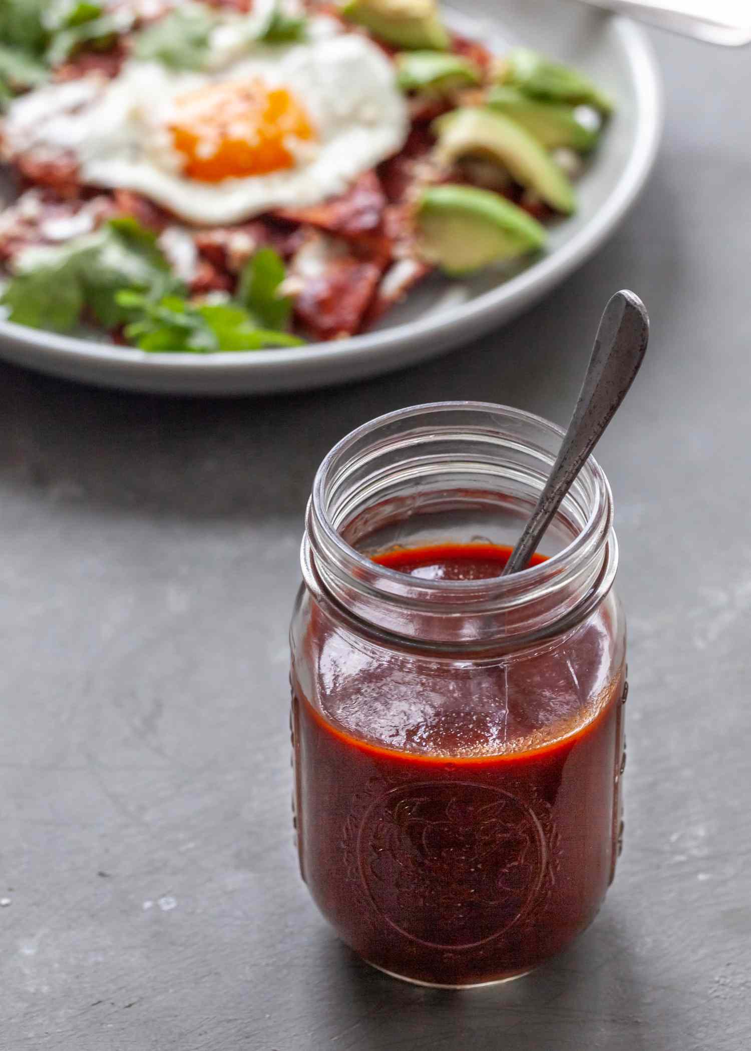A jar of chili sauce in front of a plate of chilaquiles.