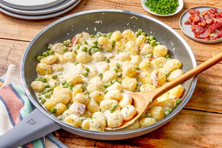Creamy Skillet Gnocchi With Bacon and Peas in a skillet with a spoon at a table setting with a stack of plates, a bowl of chopped chives, and a small plate of bacon pieces