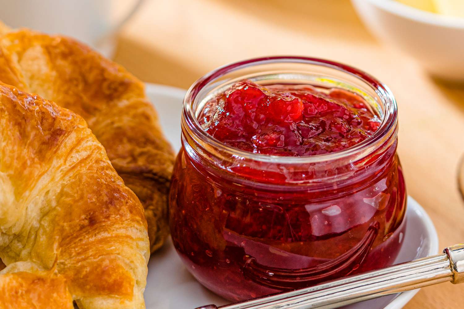 Closeup view of a small clear glass jar of strawberry jam on a plate with croissants and a knife all on a wooden tabletop