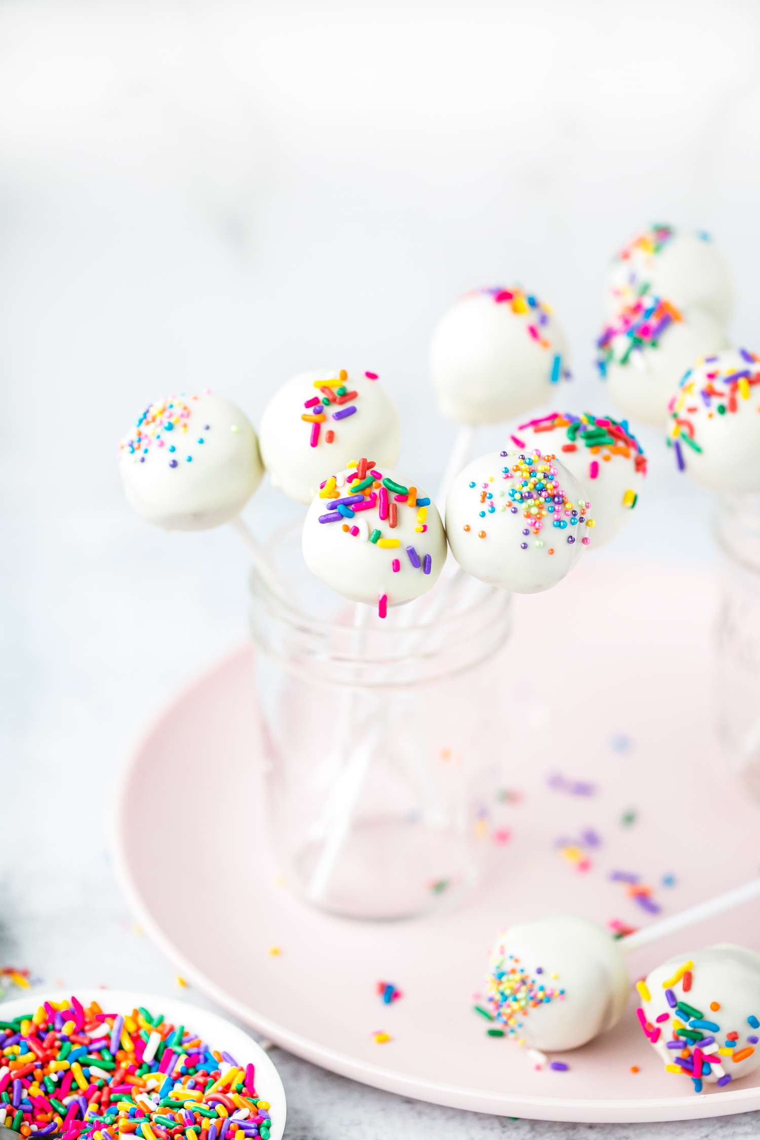 Cake Pops in Jars and Some Cake Pops on a Plate Next to Small Plate of Sprinkles