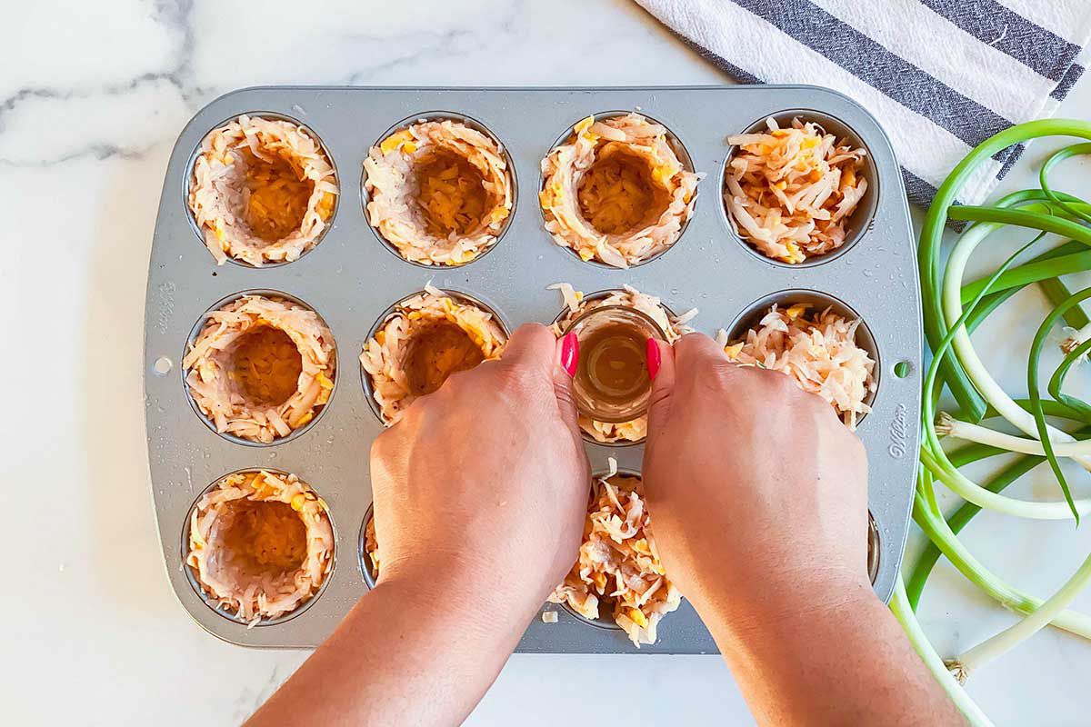 A shot glass is being pressed into the muffin tin cavity to help form the crusts for hash brown egg bites.