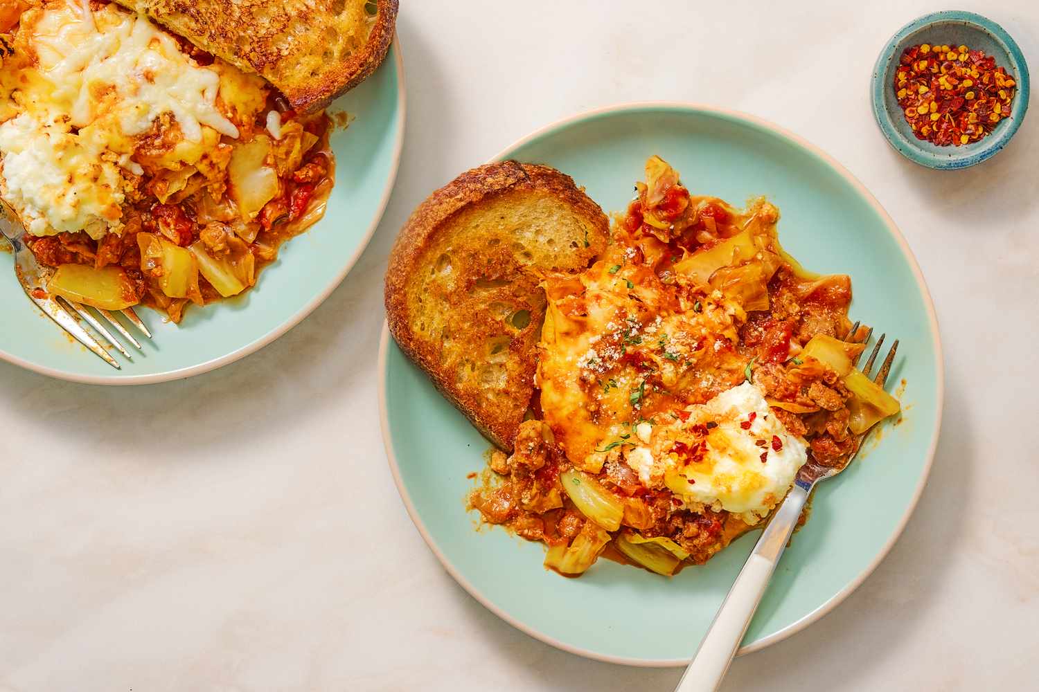 Two plates of braised cabbage lasagna with toasted bread and a side of red pepper flakes in a small bowl
