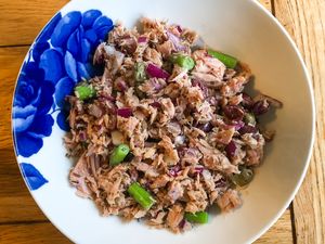 A bowl containing a mixture of tuna chopped onions green vegetables and herbs placed on a wooden table