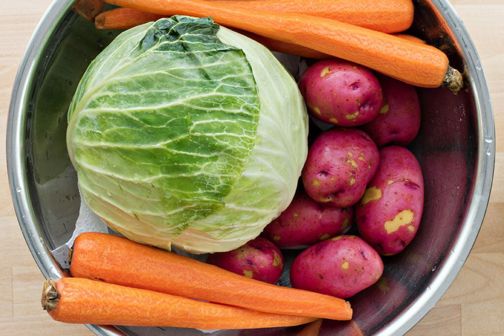 Bowl with a whole green cabbage, red new potatoes, and peeled carrots