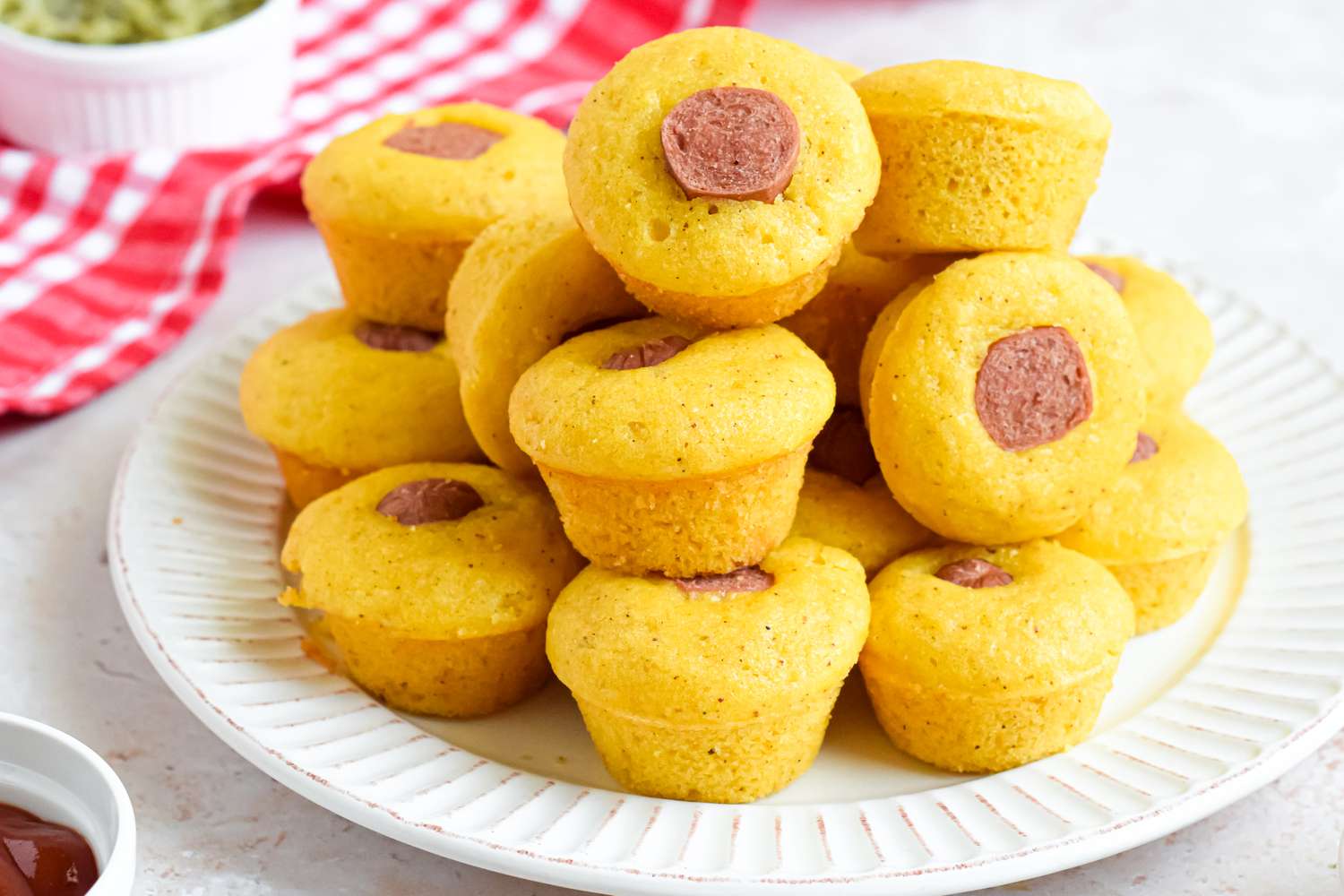 Pile of Corn Dog Muffins Stacked on a Plate, and in the Background, a Red and White Checkered Kitchen Towel and Small Bowls of Toppings (Relish and Ketchup)