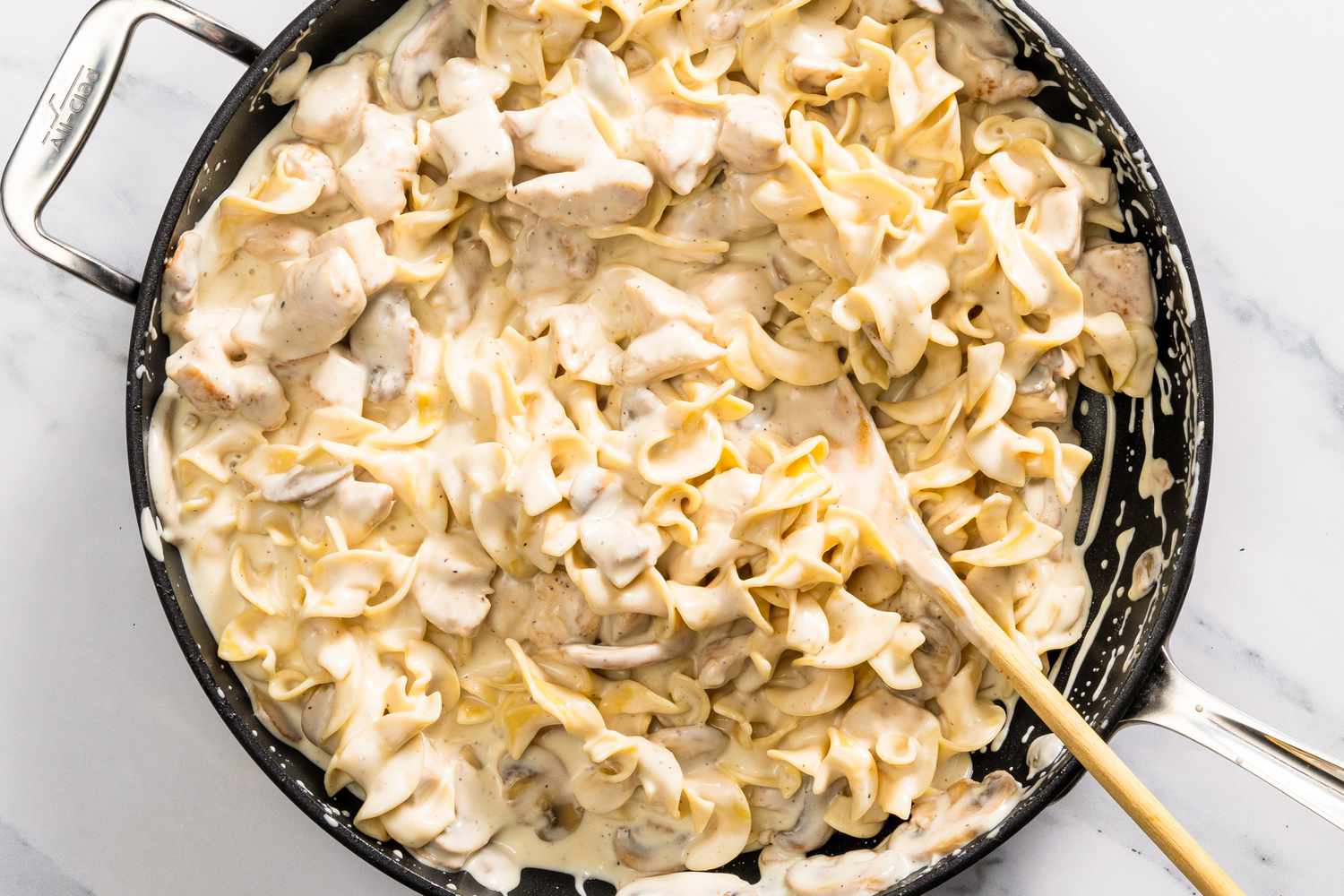 cooked egg noodles added to skillet with a wooden spoon for Church Chicken Casserole