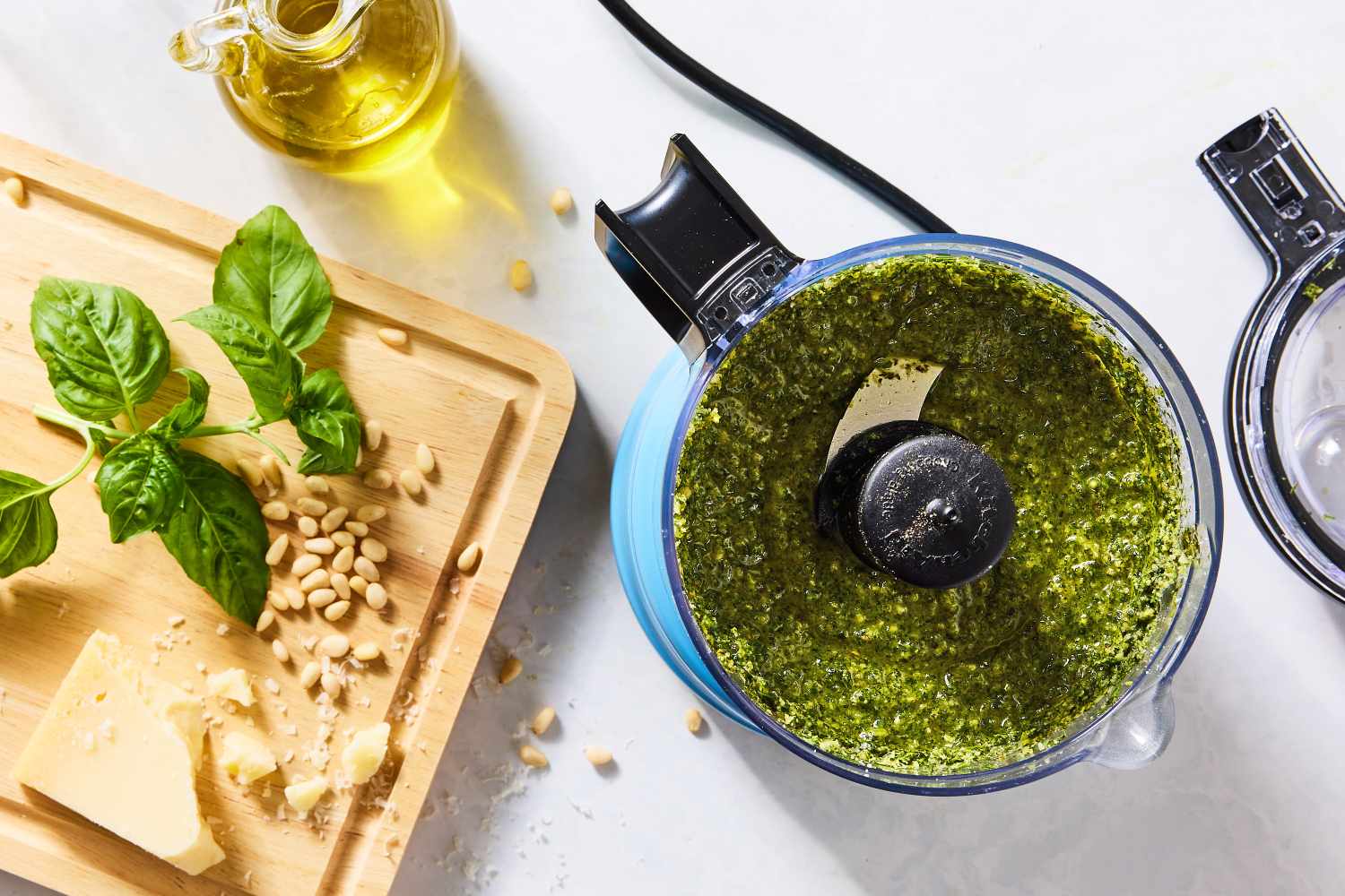 A food processor with basil pesto basil leaves Parmesan pine nuts and olive oil on a cutting board nearby