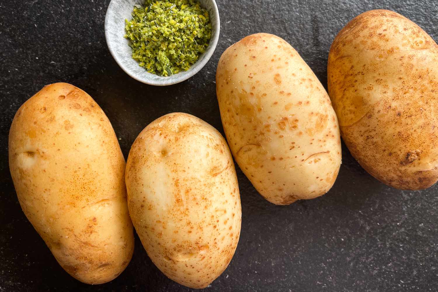 Overhead view of four uncooked potatoes and a bowl of rub on a gray countertop