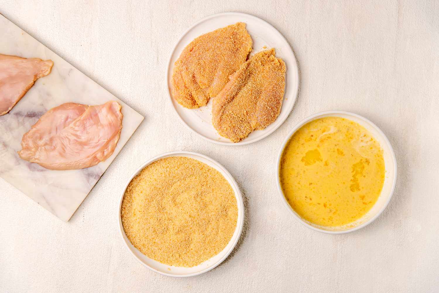 dredging set-up: flattened chicken on a tray, bowl of wet ingredients, bowl of breadcrumbs, and a plate of breaded chicken for Italian butter chicken (petti di pollo al burro) recipe