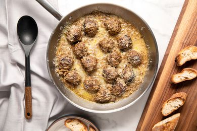 French Onion Meatballs in a Pan at a Table Setting With Toasted Baguette Slices on a Wooden Platter and a Spoon on the Counter, All on a White Table Cloth