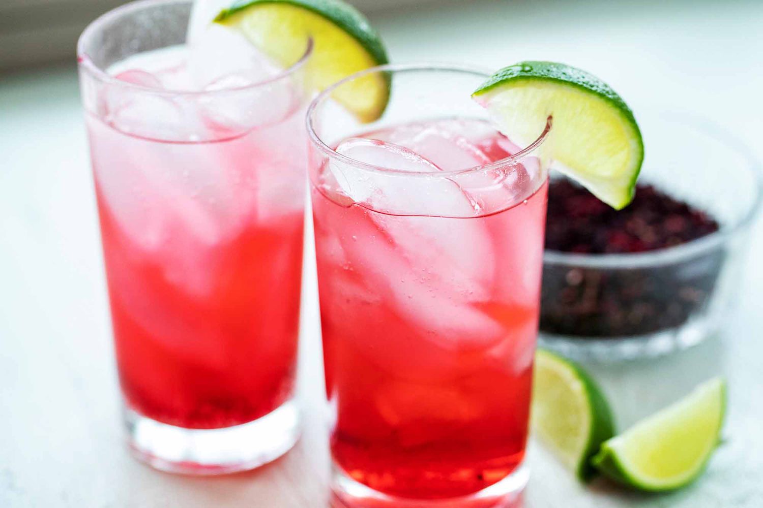 Horizontal view of two side by side tall glassed with hibiscus cocktails inside. Ice cubes are inside the glass and a lime wedge sits on the lip of each glass with additional lime wedges to the right of the glasses. Partial view of a bowl of dried hibiscus in the background to the right of the second glass.