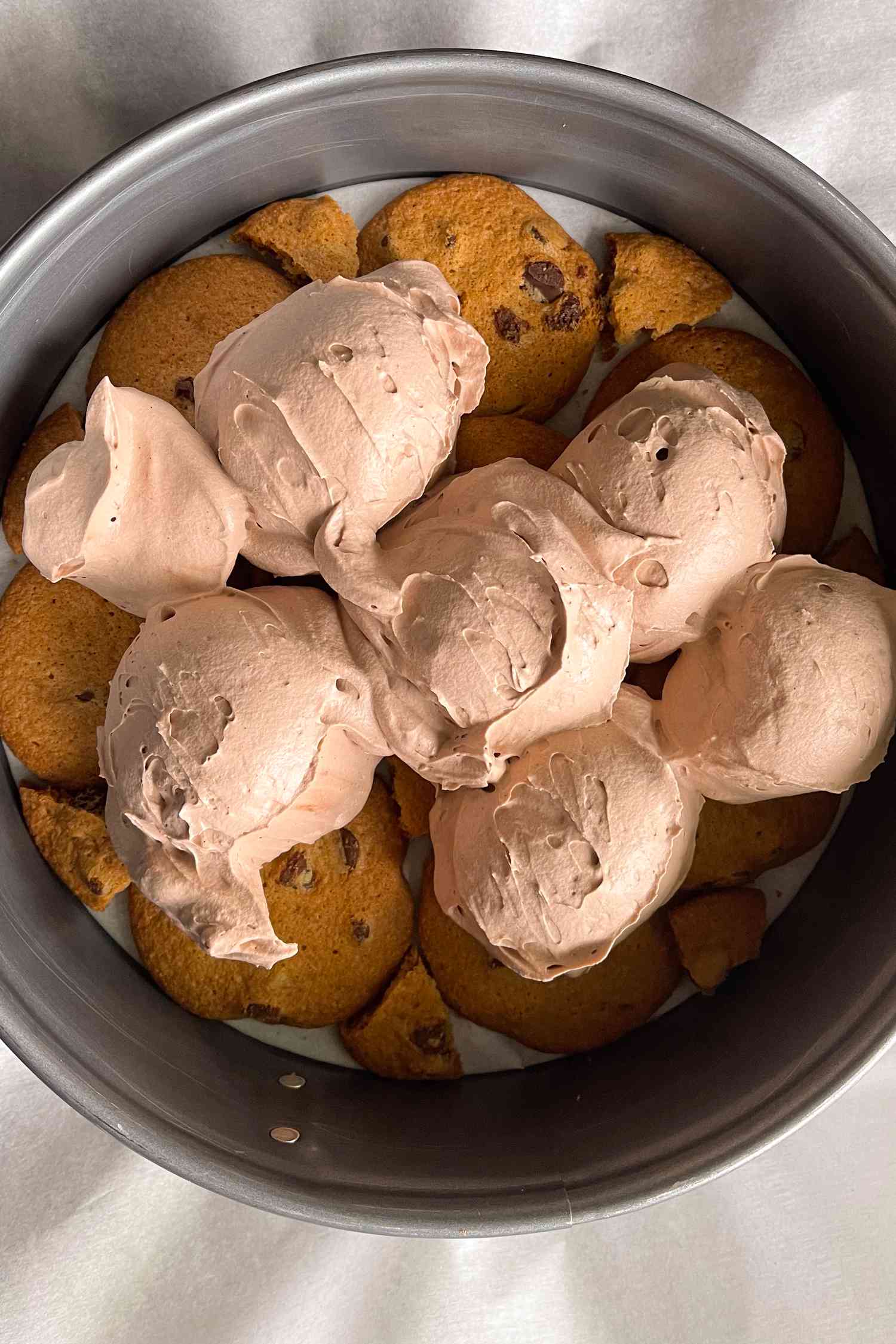 A springform pan showing a layer of chocolate chip cookies in the bottom topped with scoops of mocha cream ready to be spread into a layer