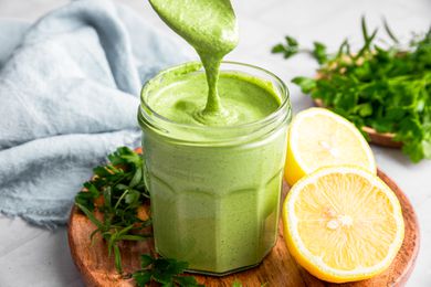 Green Goddess Dressing Dripping from a Spoon into a Jar with More Dressing. Jar is on a Wooden Plate with Herbs and Halved Lemons
