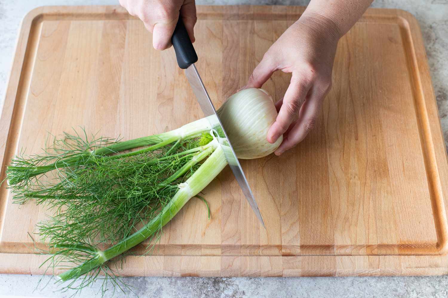 One Hand Holding the Fennel Bulb While Other Hand Cuts Off Fennel Top Using a Knife, All on a Cutting Board