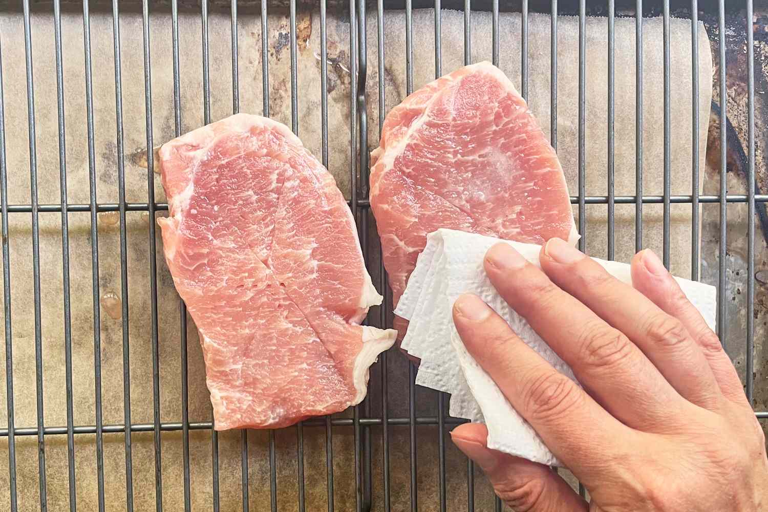 A hand using a paper towel to pat dry two pork chops on a wire rack