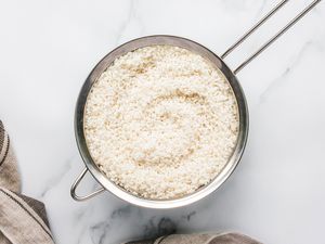 A bowl of rinsed rice sitting in a fine mesh strainer placed on a light surface with a towel nearby