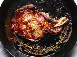 A cast iron skillet with steak cooked on the stove. Oregano sprigs are at the bottom of the pan. Oil and juices are visible in the pan.