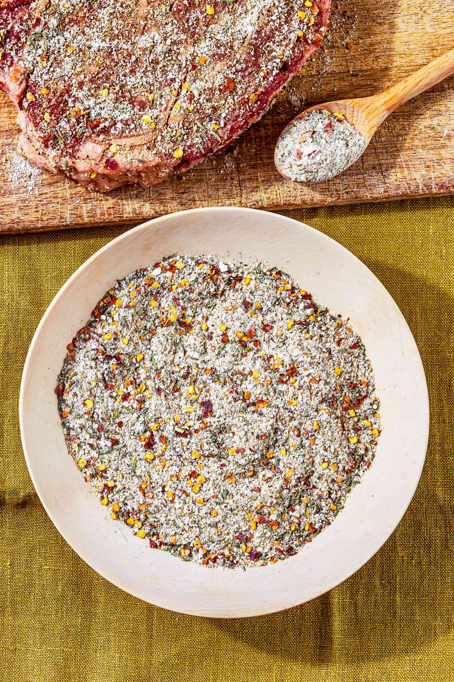 Bowl of Steak Seasoning Next to Cutting Board With a Seasoned Raw Steak and a Spoonful of Steak Seasoning