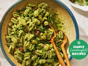 A bowl of rotini pasta with pesto sauce garnished with sundried tomatoes and accompanied by two wooden serving forks