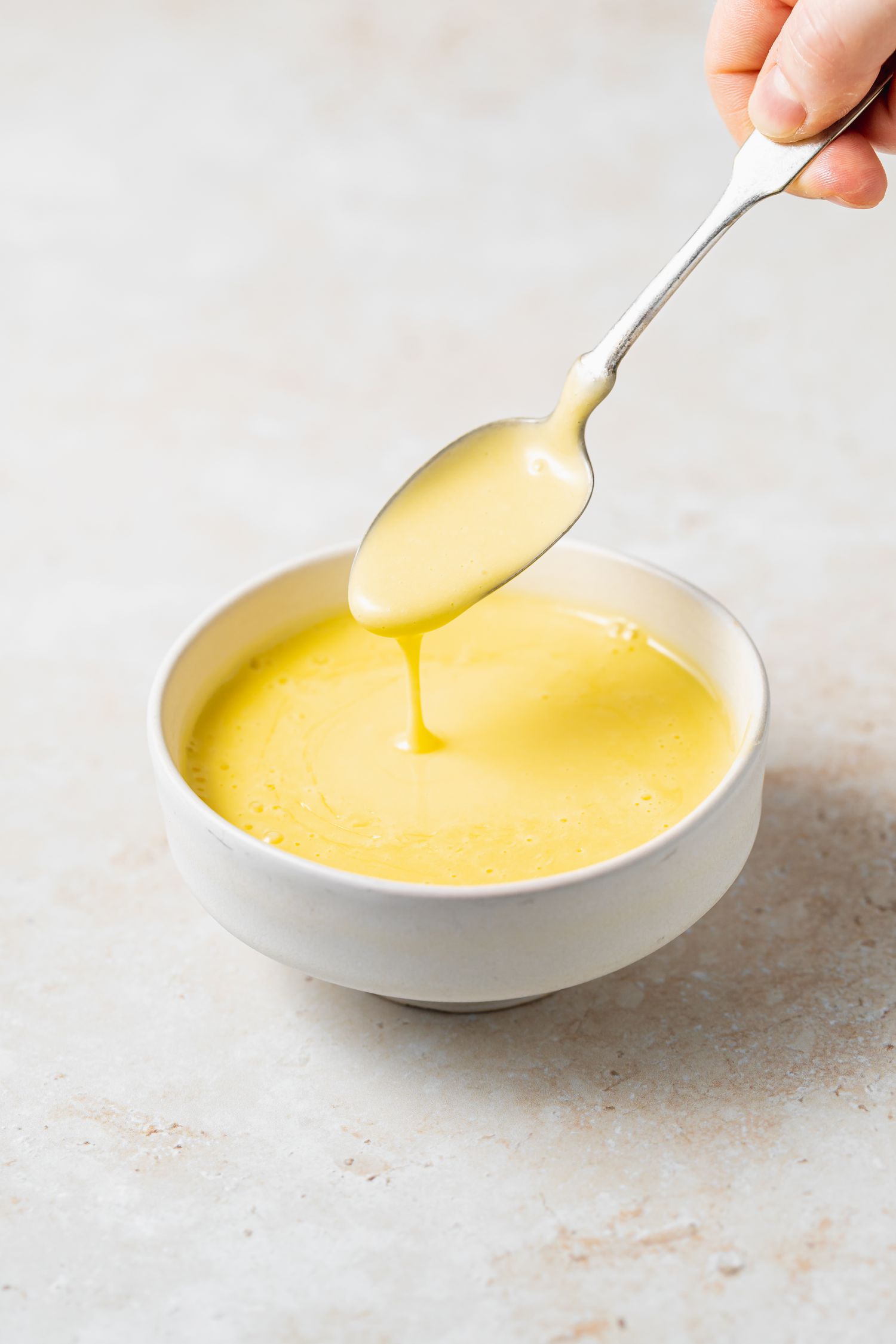 Blender Hollandaise Sauce in a white bowl with sauce dripping off of a spoon.