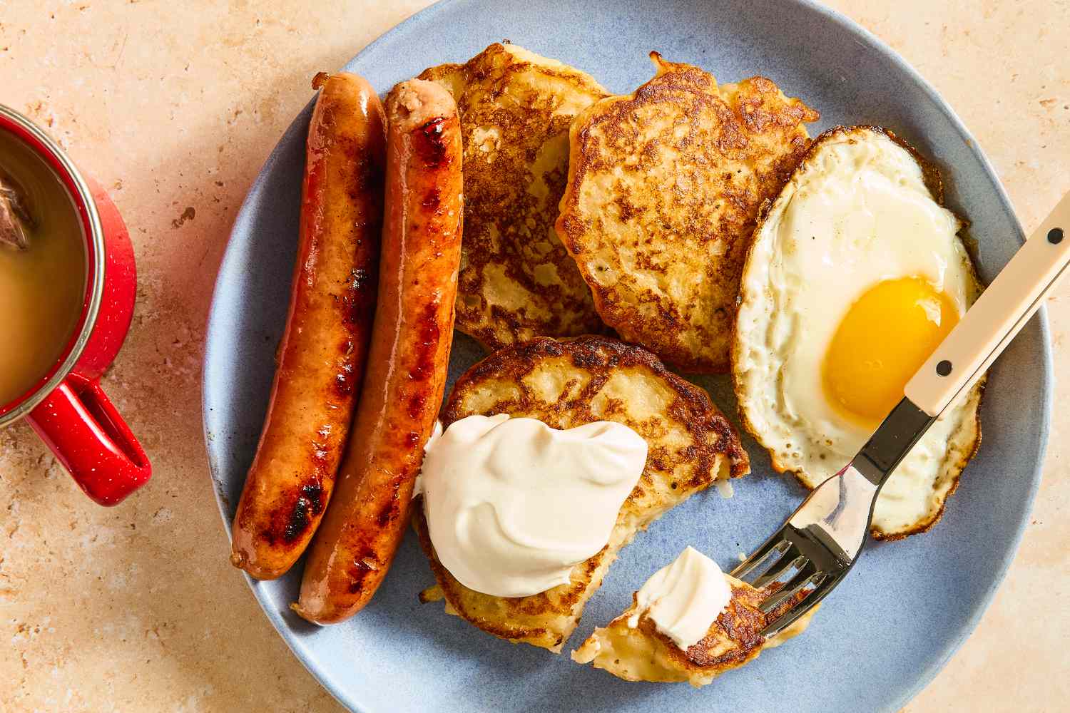 Overhead view of Irish boxty with a side of eggs and sausage next to a fork with a bite on it on a plate, all next to a coffee mug on a stone countertop