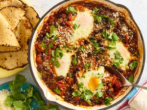 A round baking dish with the black bean and egg bake recipe, next to a plate of blistered corn tortillas and garnish of cilantro and fresh cheese