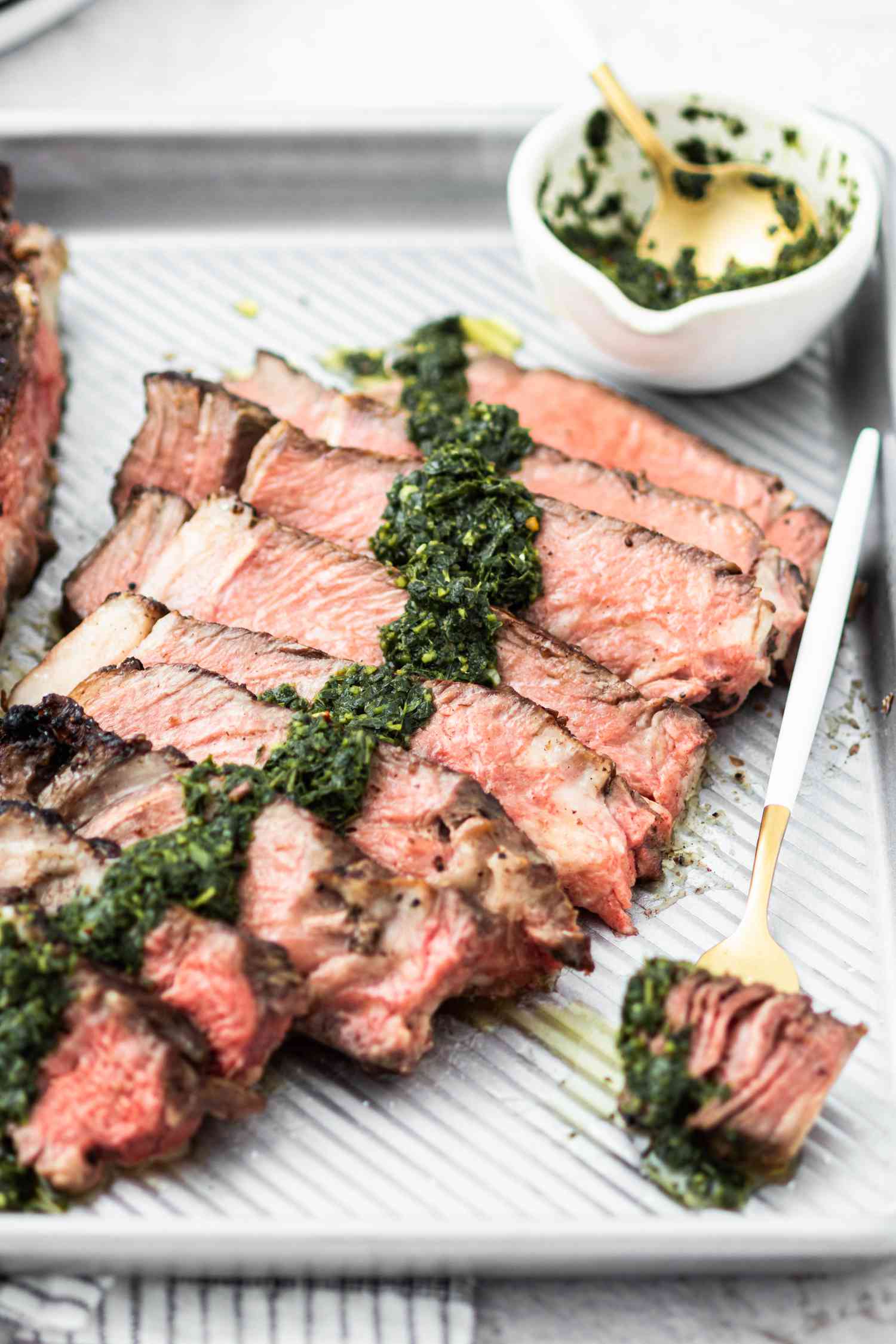 A fork holding a piece of cowboy ribeye steak set next to more slices on a baking sheet.