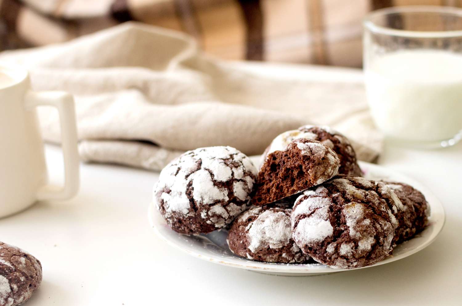 A plate of chocolate crinkle cookies with a cup of milk and a white cup nearby on a table setup