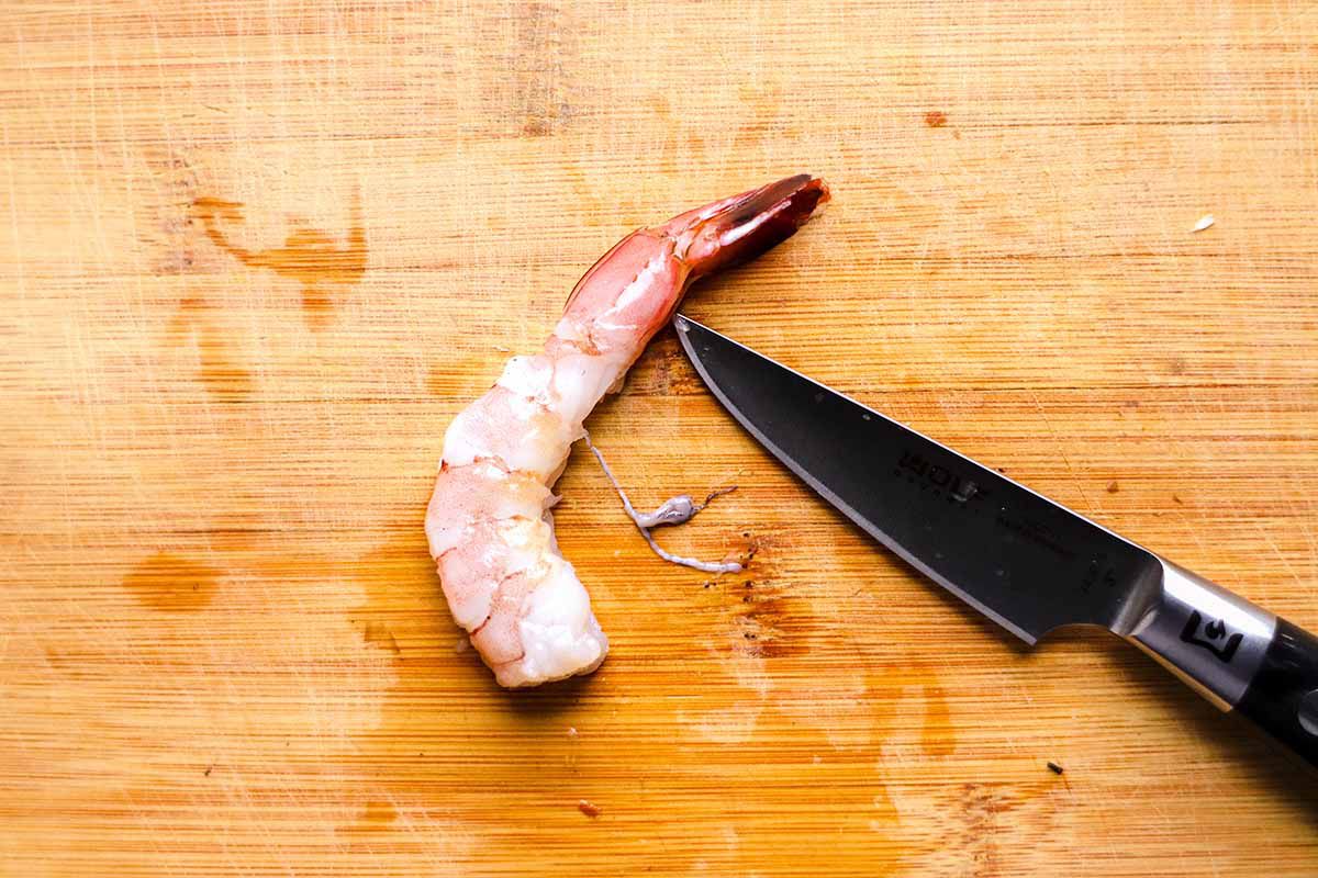A raw shrimp is on a cutting board with a knife blade cutting the vein out of it.