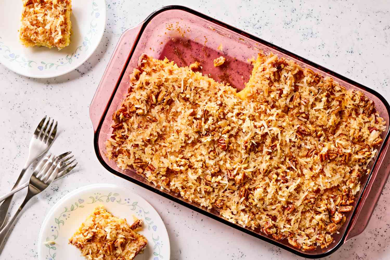 Overhead view of a glass baking dish of pineapple coconut cake with two slices removed next to two small plates with slices along with three forks