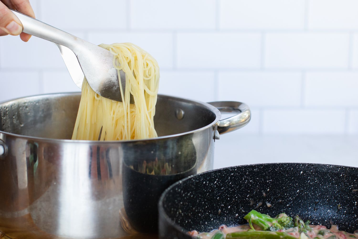 Tongs with cooked spaghetti noodles in a pot with a the pasta primavera vegetables next to it in a skillet.