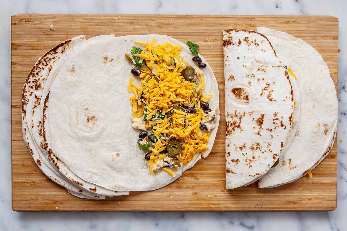 Healthy breakfast quesadillas on a wooden cutting board. One tortilla has shredded cheese, scrambled eggs and black bean filling on half. To the right two quesadillas have already been filled and folded.
