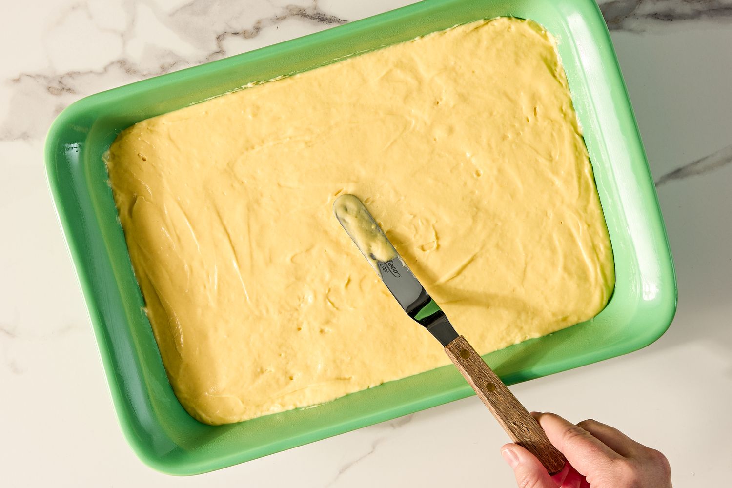 A hand uses a spatula to spread cake batter in a green baking dish